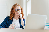 Horizontal shot of pleasant looking successful professional female lawyer learns clients case, works on modern laptop computer, dressed in formal apparel and transparent glasses, poses in office