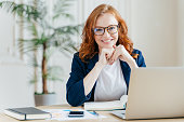 Pleased cheerful red haird female economist develops financial startup project, poses in office interior, works in business sphere, dressed in formal clothes, has happy expression, owns company