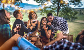 Young friends having fun at vineyard after sunset - Happy people millennial camping at open air pic nic under bulb lights - Youth friendship concept with guys and girls drinking wine at barbeque party