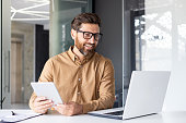 Portrait of a young architect, designer working in the office at a laptop, talking on a video call, holding a tablet in his hands