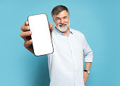 Handsome Excited Mature Man Showing Pointing At Empty Smartphone Screen Posing Over Light Blue Studio Background, Smiling To Camera. Check This Out, Cellphone Display Mock Up